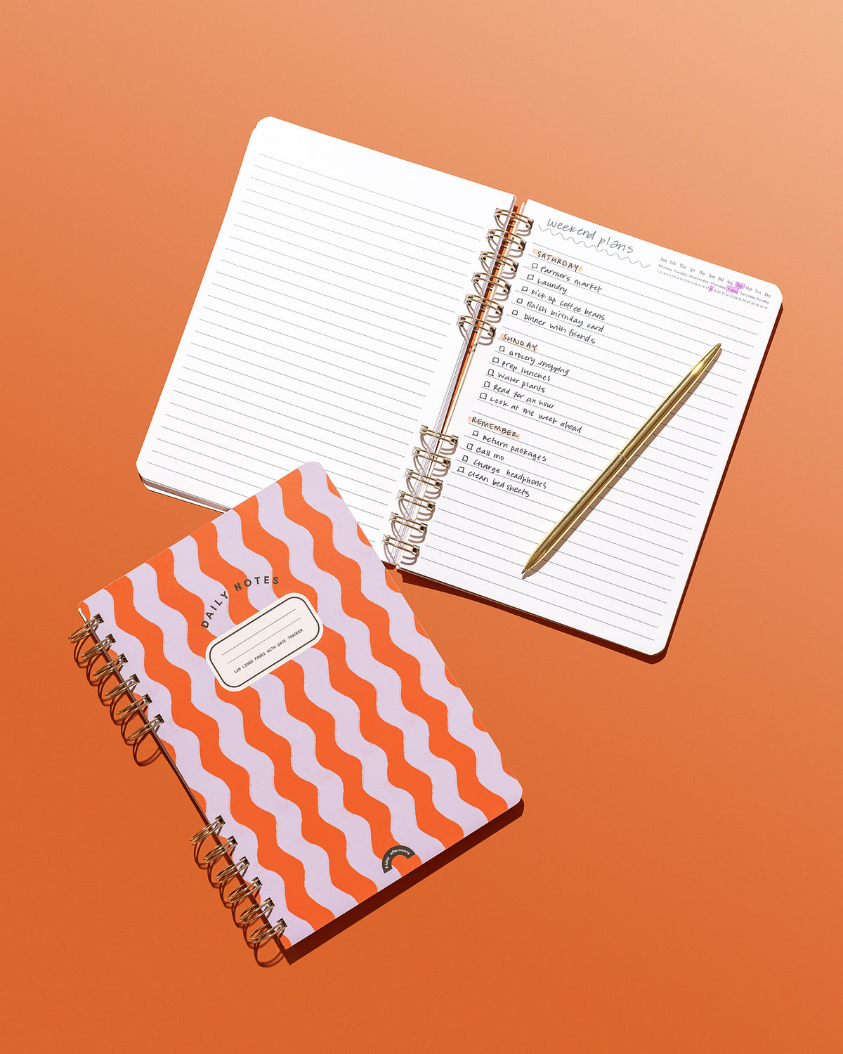 Poppy & Lavender Swirls Daily Notes notebook beside an open lined notebook with handwritten weekend plans and a gold pen on an orange background.