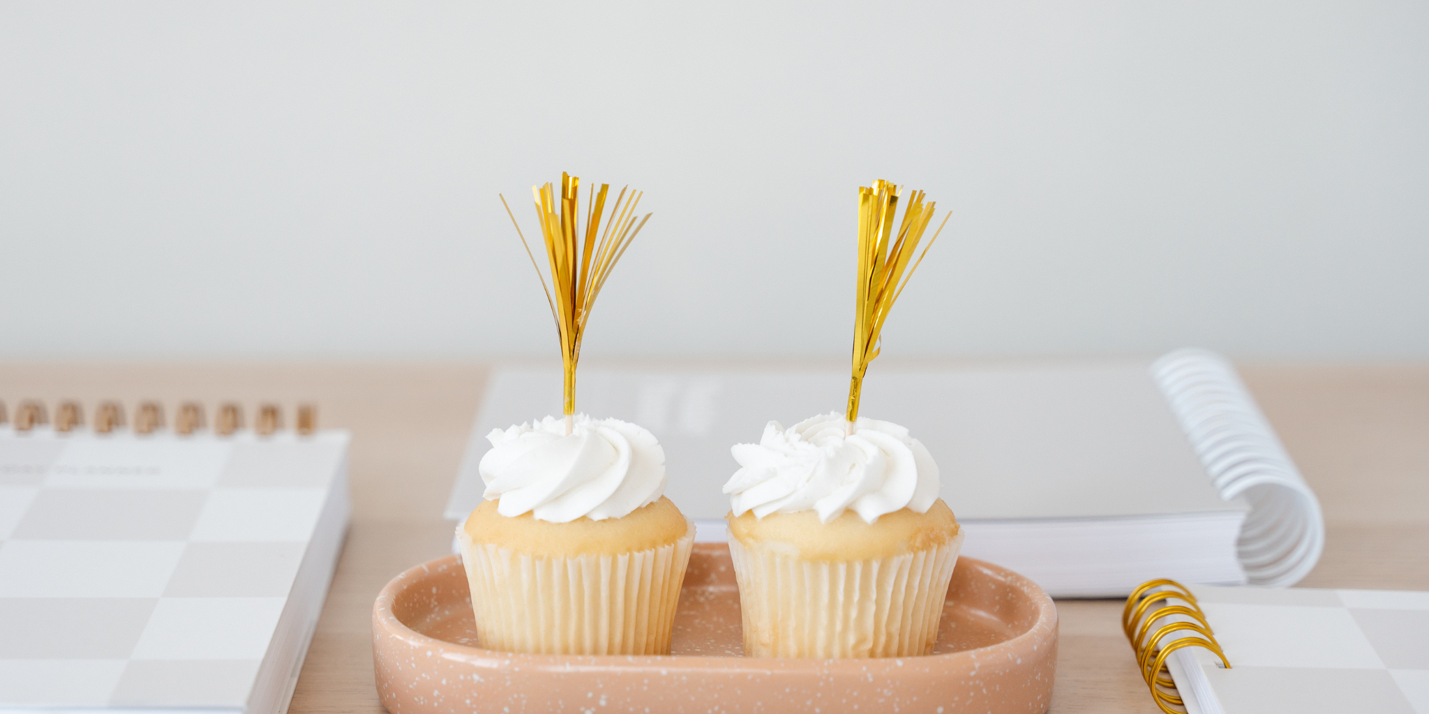 Two cupcakes with white frosting and gold picks on a pink plate, placed on a light surface.