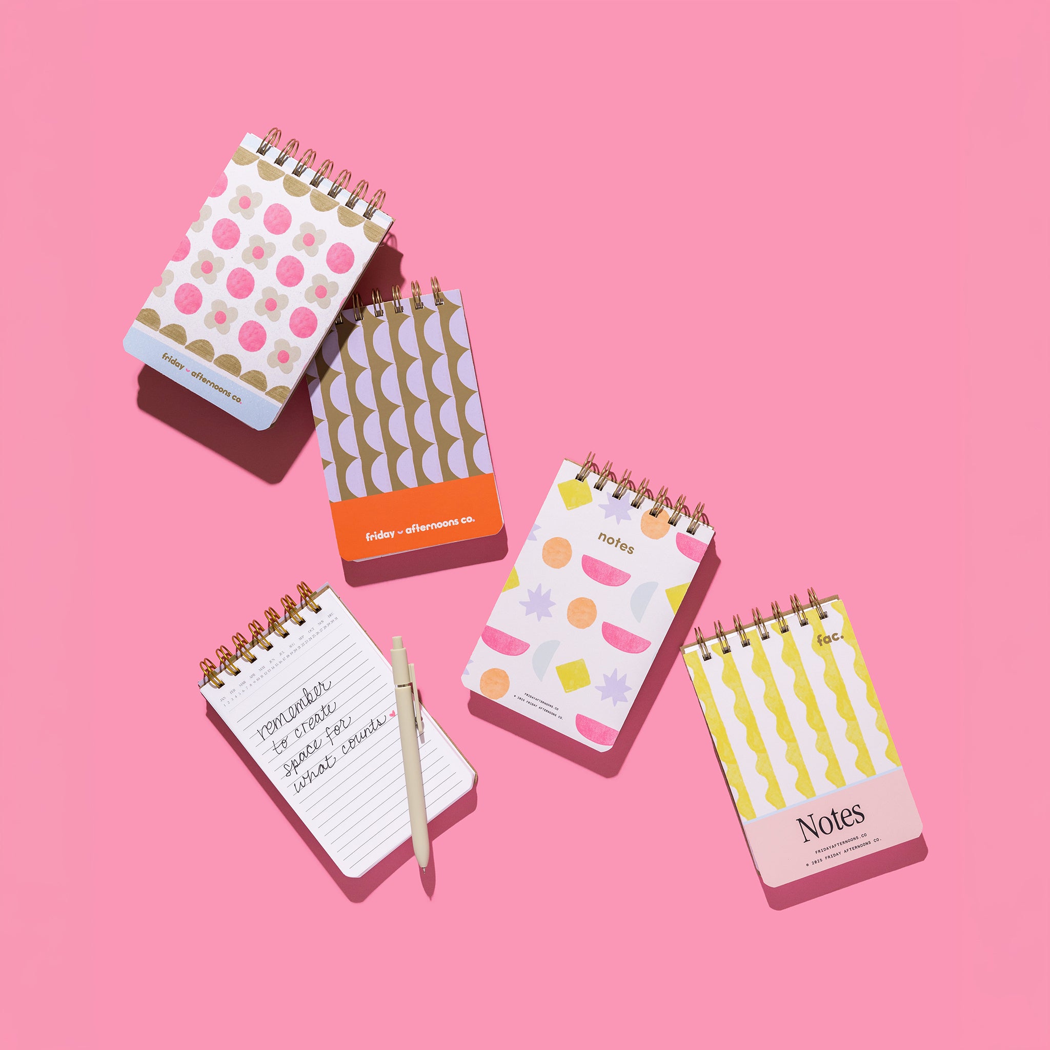 Mini Memo Notebook collection in four cover designs shown on a pink background with one notebook open, lined pages, and a pen.