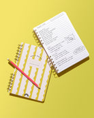 Lima Wavy Stripes Daily Notes notebook beside open lined pages with a handwritten daily plan and pink pencil on a yellow background.