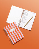 Poppy & Lavender Swirls Daily Notes notebook beside an open lined notebook with handwritten weekend plans and a gold pen on an orange background.