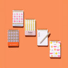 Mini Memo Notebook collection in four cover designs shown on an orange background with one open lined notebook and white pen.