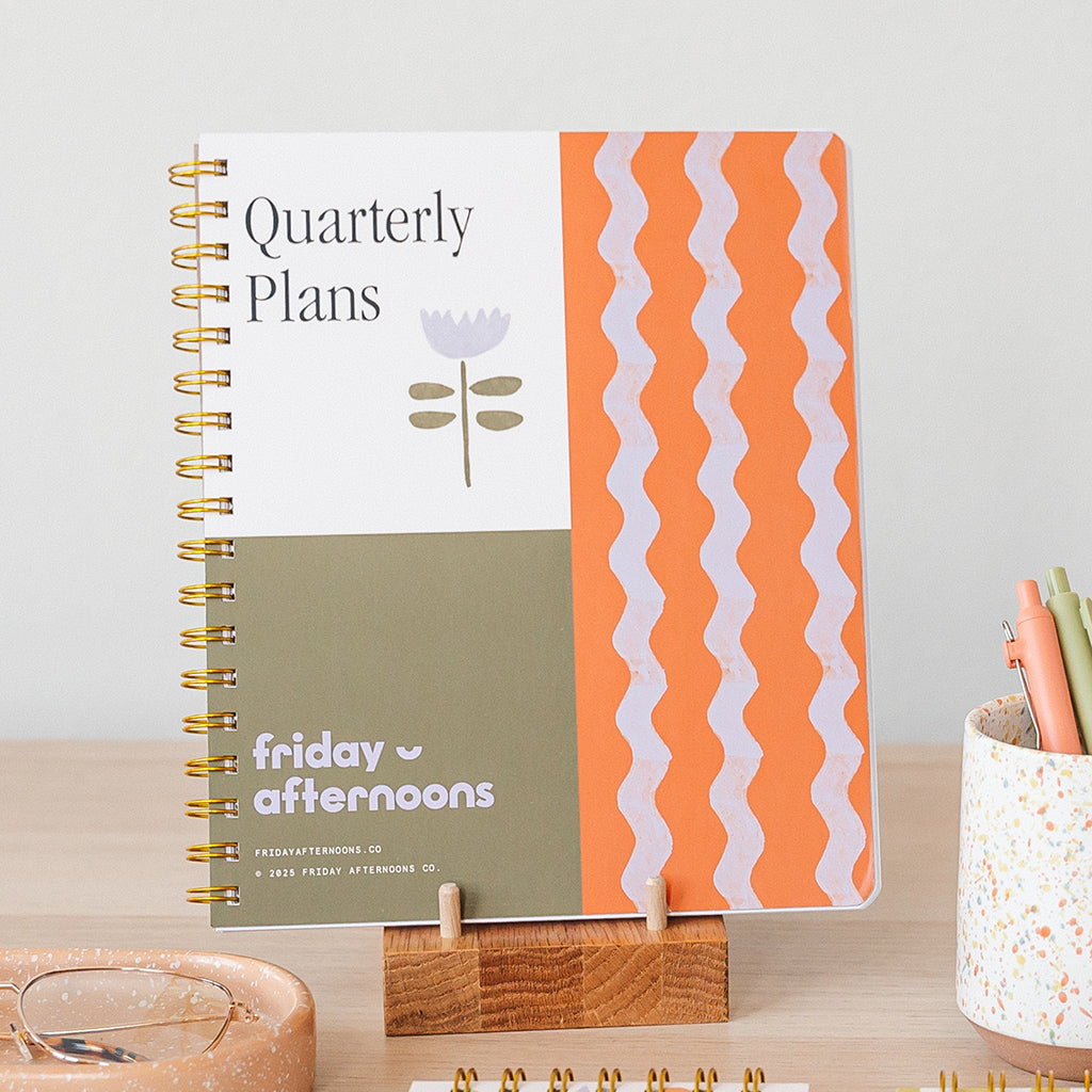 Quarterly Plans Planner standing on a wooden stand with an orange and olive graphic cover from Friday Afternoons Co.