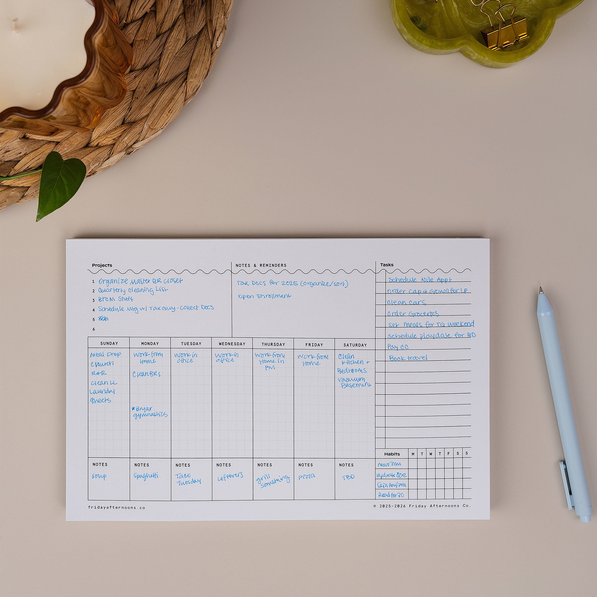 Task Track Pad weekly planner notepad filled out with handwritten tasks, shown on a desk with a pen.