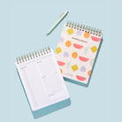 Bright Shapes Workday Planner with an open daily planning page, top-spiral binding, and pen on a light blue background.