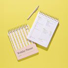 Lima Wavy Stripes Workday Planner styled on a bright yellow background, shown closed with the yellow wavy stripe cover and pale pink title panel beside an open daily planning page, with a white pen above.